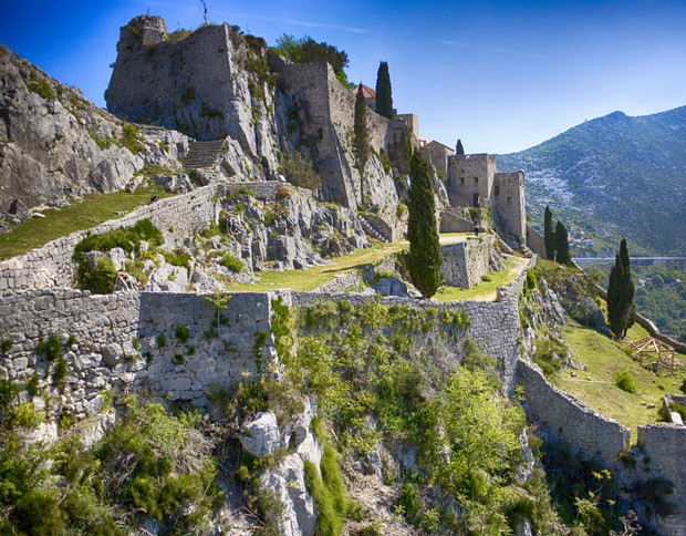 Ancient stone fortress perched on a rocky hillside, surrounded by lush green vegetation. Blue sky and distant mountains in the background create a picturesque, historic landscape.